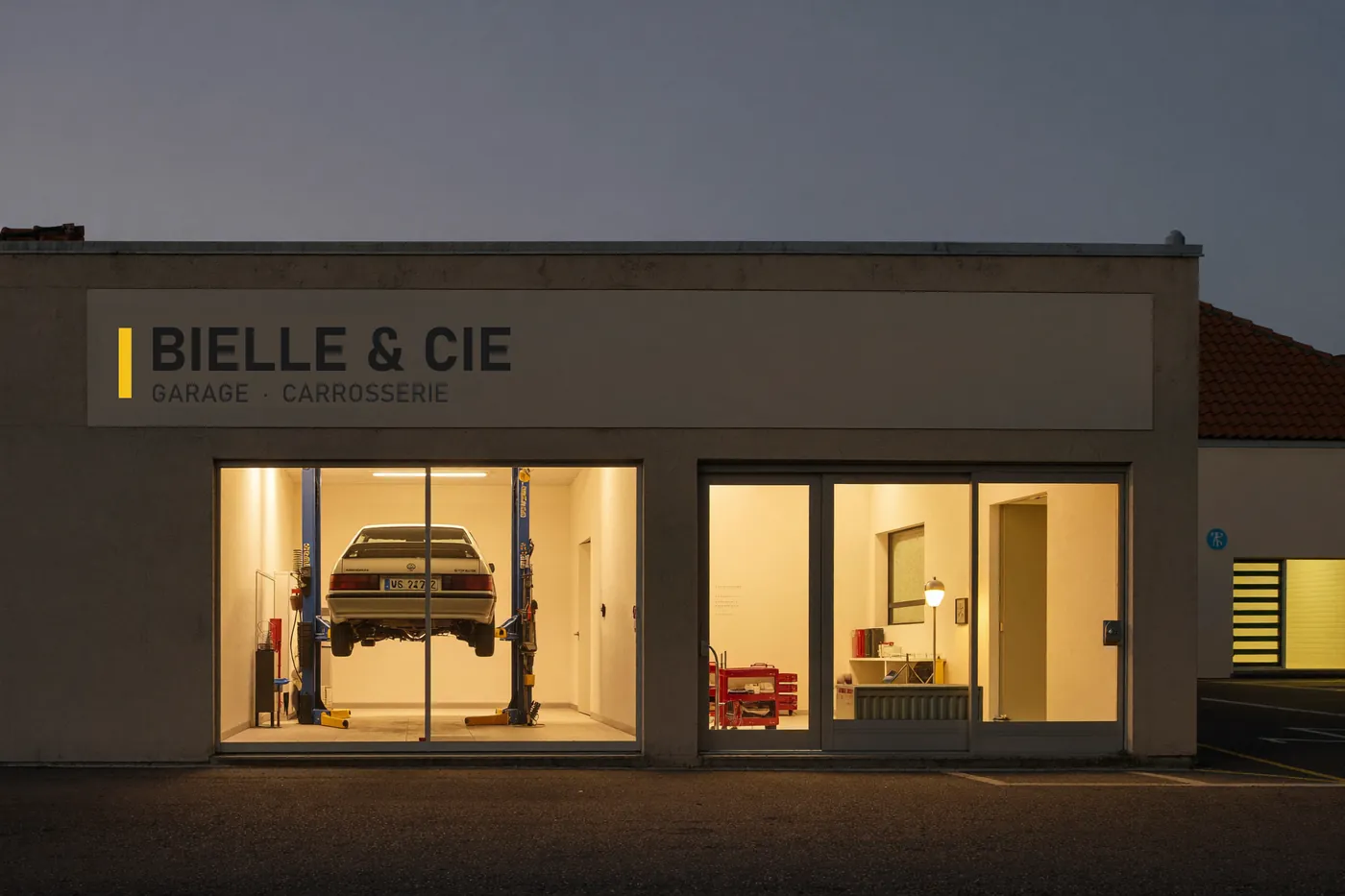 La façade du garage Bielle & Cie à la tombée de la nuit, portes ouvertes et éclairées